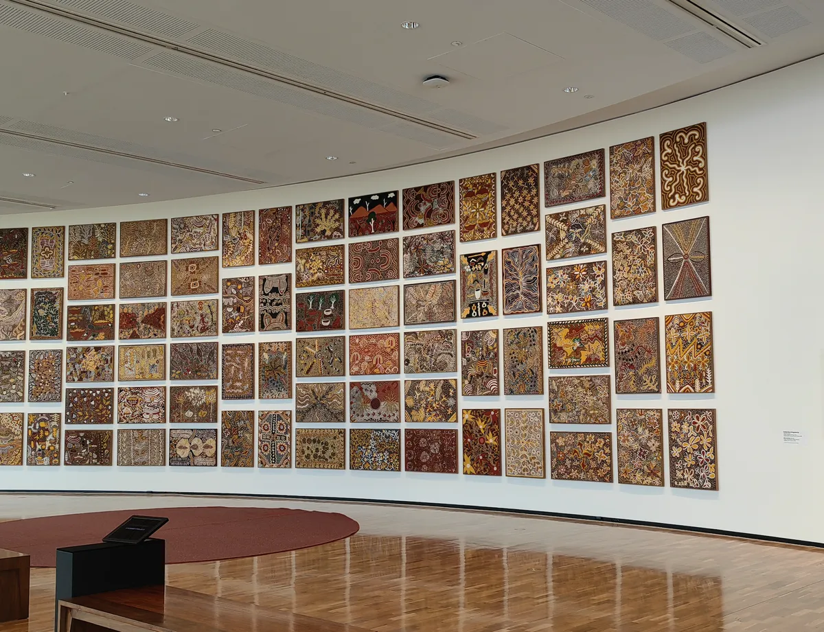 A curved gallery wall salon-hung with dozens of small Aboriginal artworks in a grid, National Gallery of Australia