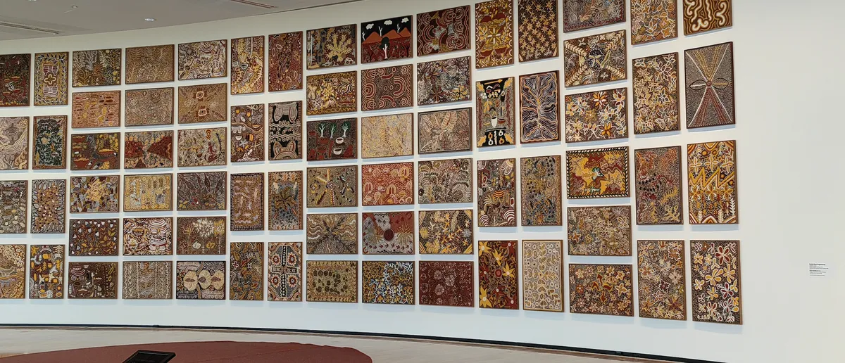 Wide view of the same curved salon-hung gallery wall of small Aboriginal artworks, National Gallery of Australia